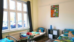 A dormitory room with bunk beds, a radiator, a window with curtains, a desk with a chair, and a colorful artwork on the wall.