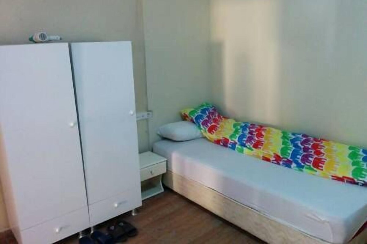 Une petite chambre avec un lit simple, une armoire blanche, une table de chevet et une couvre-lit aux couleurs vives.