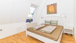 A bedroom with a sloped ceiling, a bed with striped bedding, a nightstand, a wall-mounted lamp, and a window with a view of buildings. - Vienna Hotspot - Rathaus (Vienna)