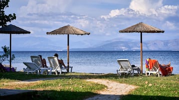 Eine Strandbucht mit Liegestühlen und strohgedeckten Sonnenschirmen, mit Blick auf ein ruhiges Meer und entfernte Berge.