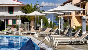 A poolside area with lounge chairs and umbrellas, a clear sky, and a building with a red and white facade. - Fardini Seaside Hotel (Corfu)