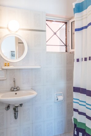 A bathroom with a white sink, a mirror, a window with a metal grid, and a striped shower curtain. - Fardini Seaside Hotel (Corfu)