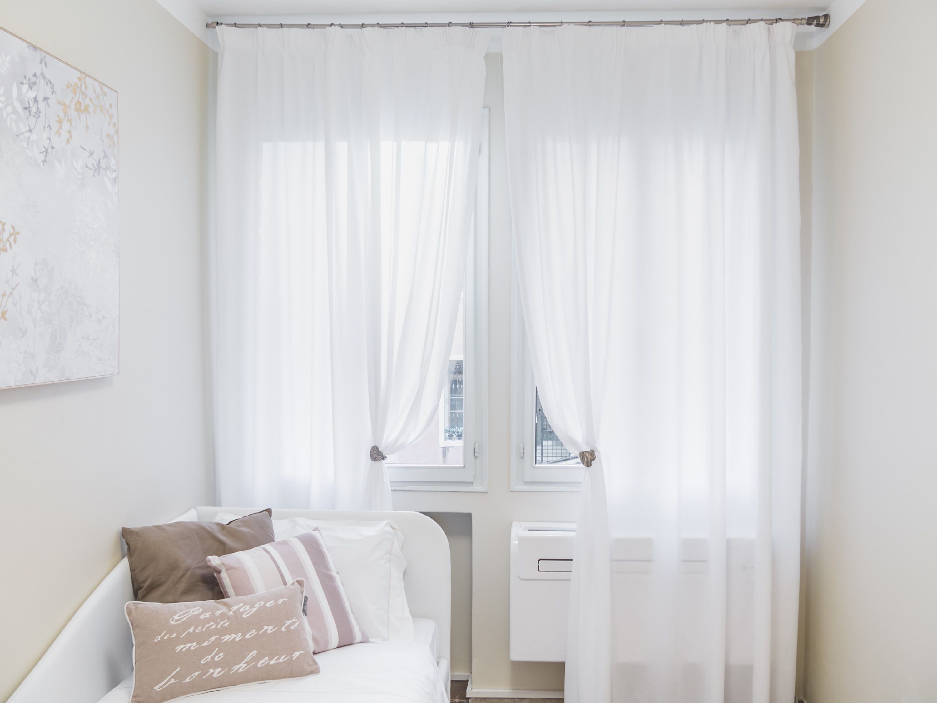 Un dormitorio con cama, cortinas blancas, un radiador y una obra de arte colgada en la pared.