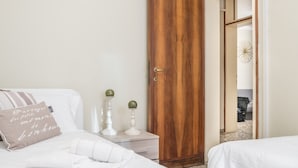 Un dormitorio con puerta de madera, cama con ropa de cama blanca, mesita de noche con dos velas y un cojín decorativo.