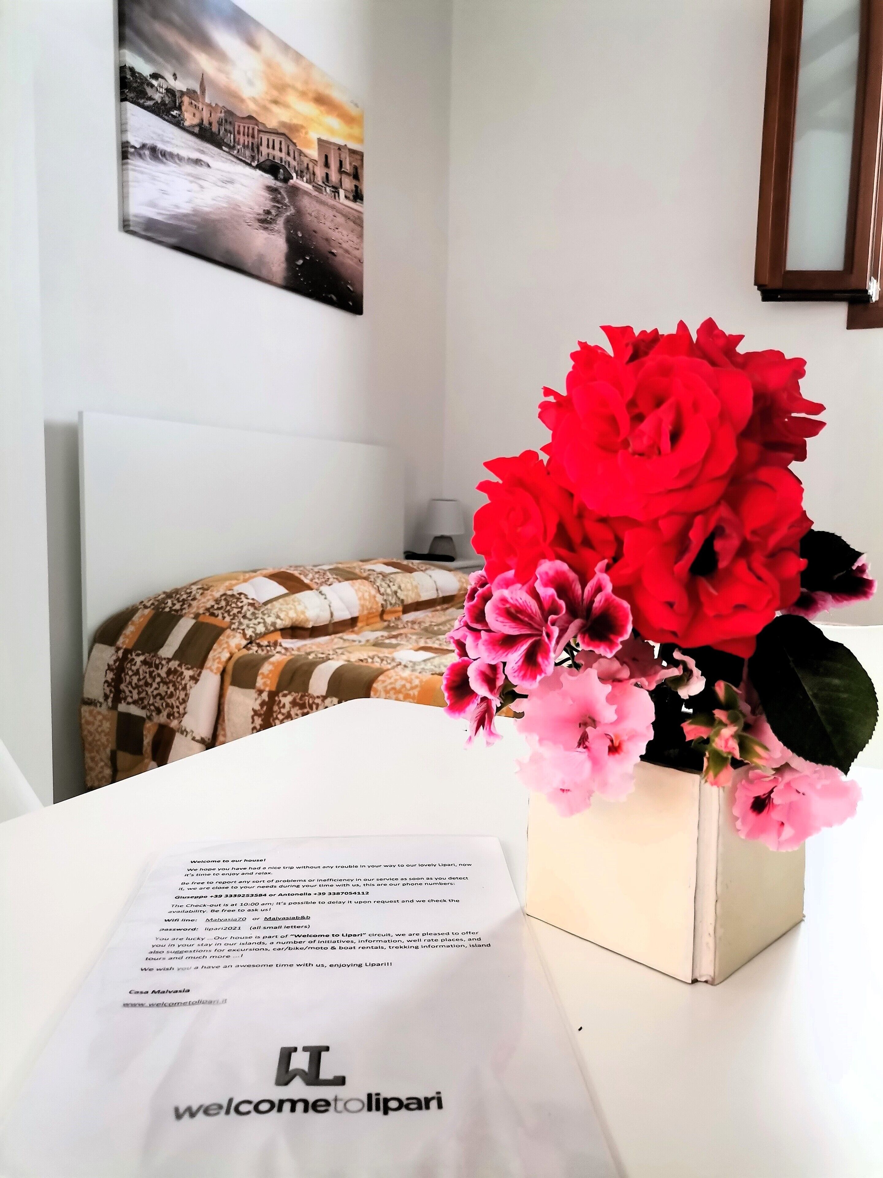 A room with a bed, a table with a welcome card, and a vase with flowers.