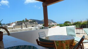 Una terraza con una mesa y sillas, con vistas a un pueblo costero y edificios, y una colina al fondo.