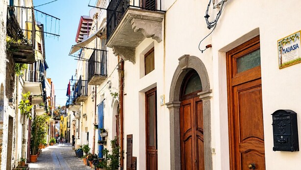 Una estrecha calle adoquinada flanqueada por edificios tradicionales, con balcones adornados con plantas en macetas, puertas de madera y un cartel que indica un lugar.
