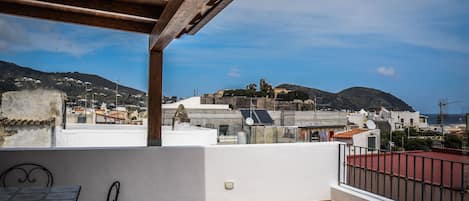 A balcony with a tiled floor, a table with chairs, and a view of a coastal town with buildings and hills in the background.