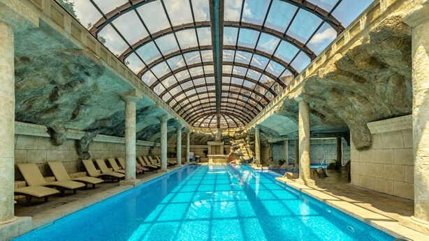 Una piscina interna con soffitto in vetro, pareti in pietra e poltroncine relax lungo il bordo.