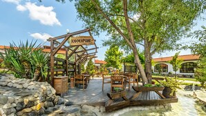 A wooden pergola with a sign reading "КАКАПЛИК" in a garden area with stone flooring, wooden benches, and tables.
