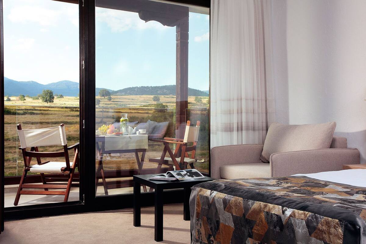 A room with a bed, a sofa, a table, and a chair. There is a view of a landscape through a large window.