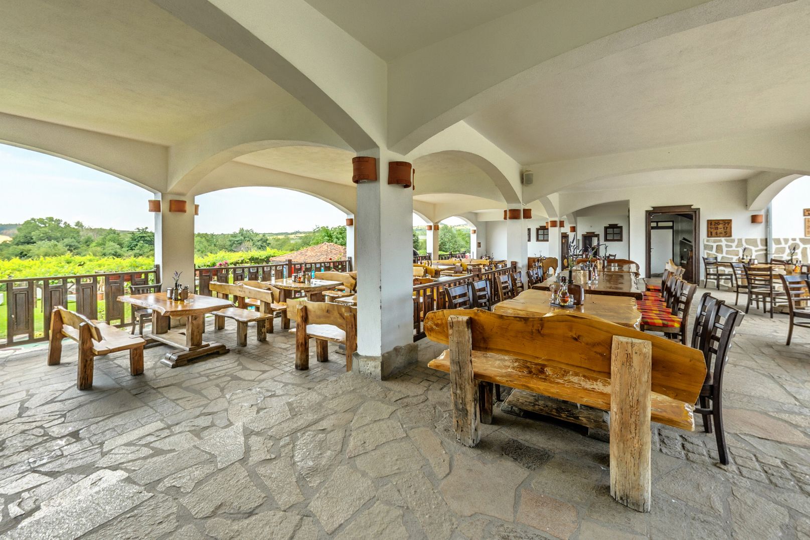 A spacious outdoor dining area with stone flooring and wooden furniture, featuring a view of a vineyard.