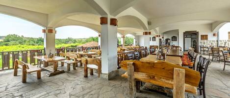 A spacious outdoor dining area with stone flooring and wooden furniture, featuring a view of a vineyard.