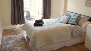 Un lit bien fait, avec une couette matelassée, une table de chevet et une chaise, dans une pièce au plancher en bois et ornée d’un petit tapis.