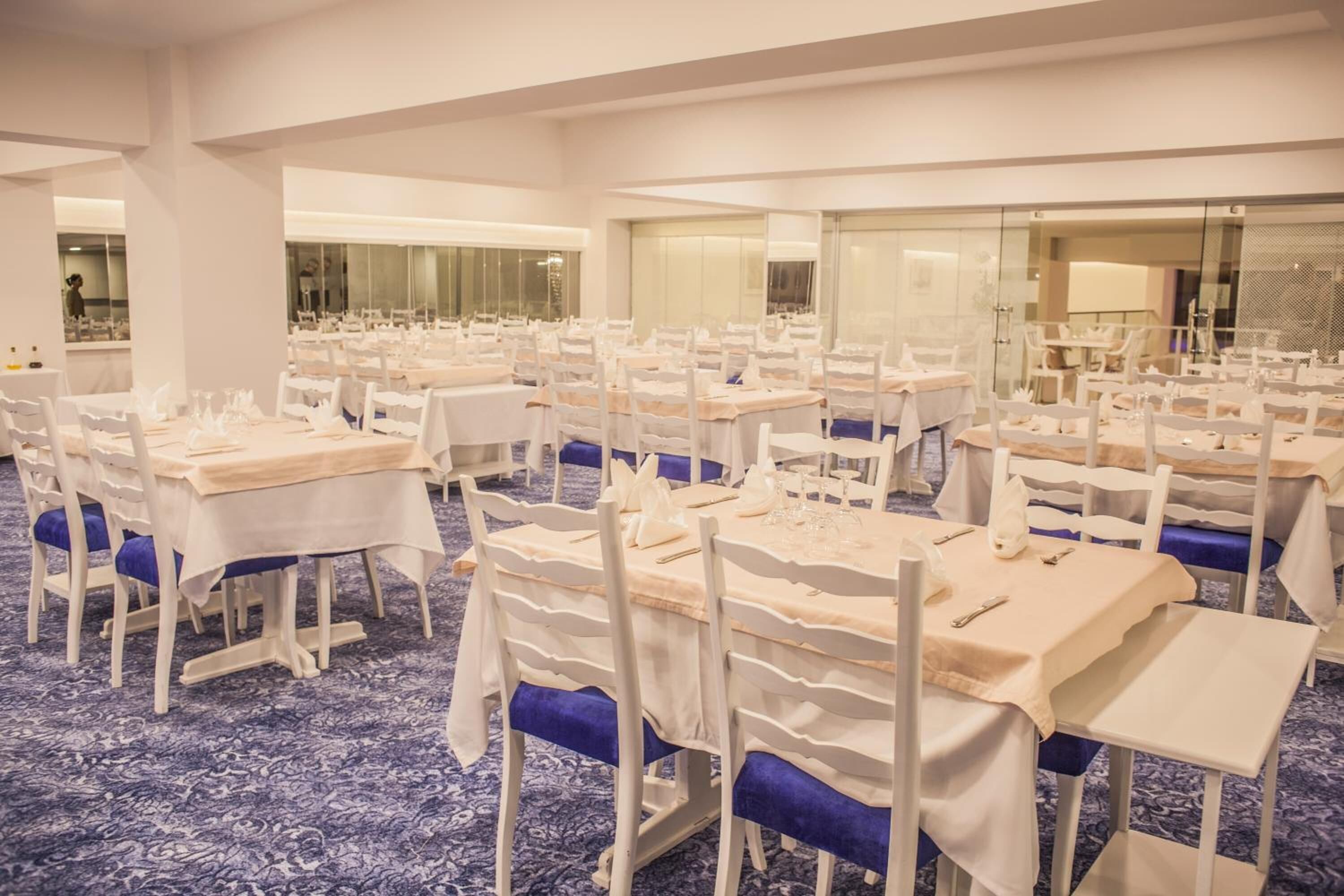 Un salón de banquetes con varias mesas dispuestas en filas, cada una con manteles blancos y sillas azules.
