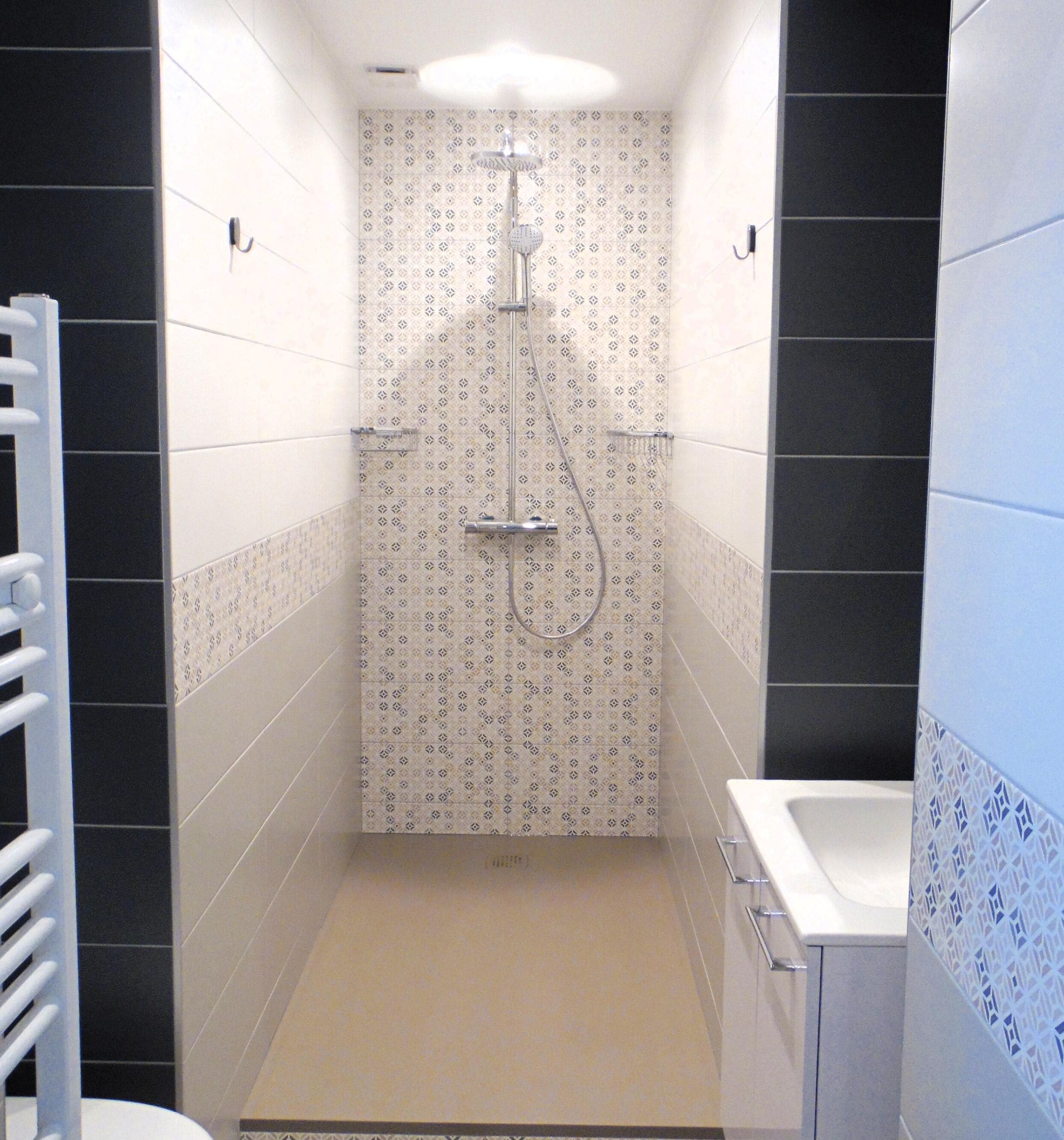 A narrow shower stall with a patterned wall, a handheld showerhead, and a glass-enclosed sink.