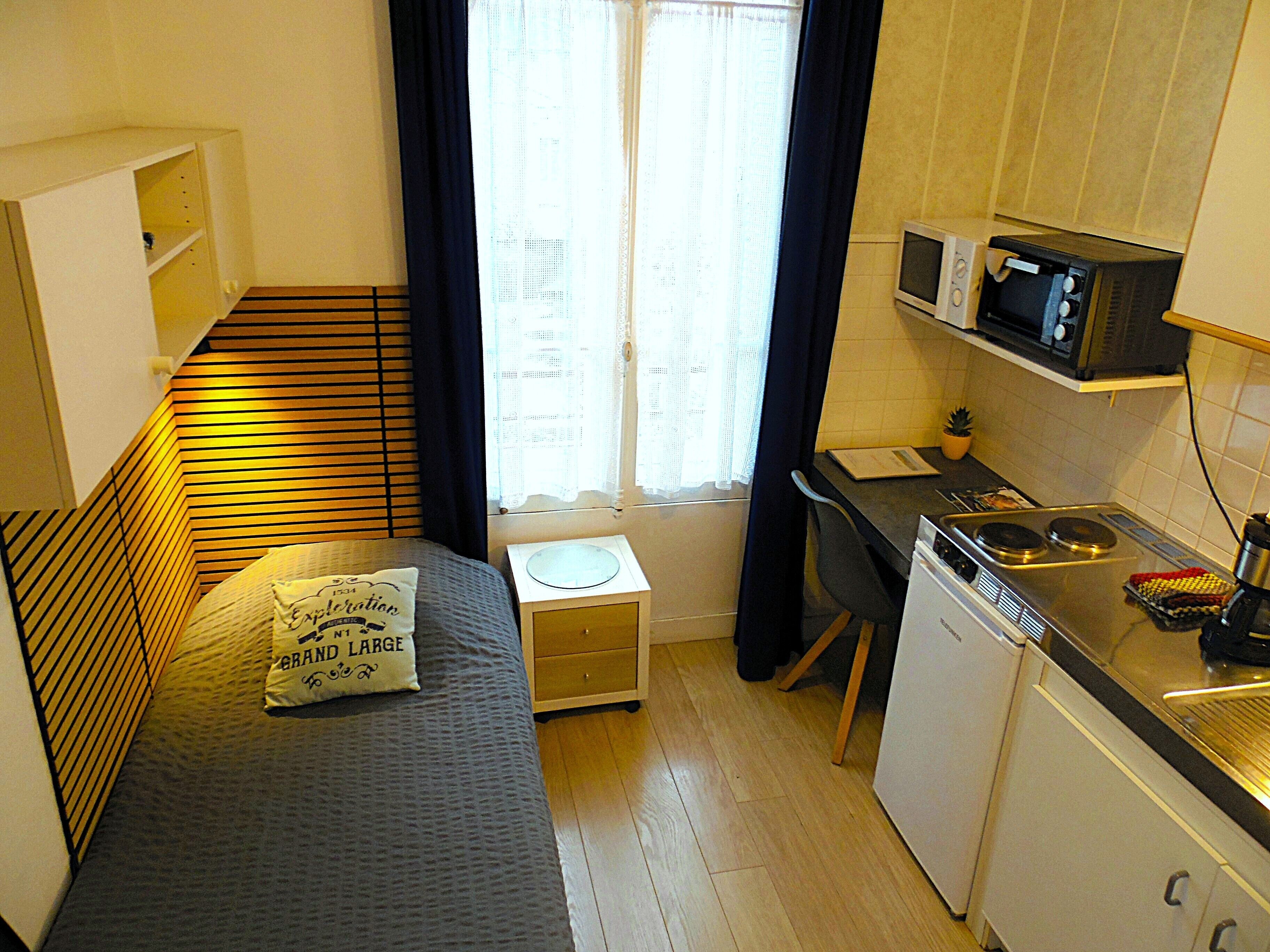 A compact room with a bed, a small bedside table, a microwave, a sink, and a window with curtains.