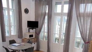 Een kamer met een eettafel, stoelen, een televisie, een magnetron en uitzicht op gebouwen door grote ramen.