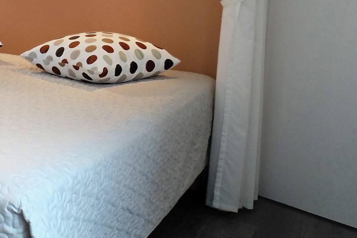En säng med ett vitt täcke och en kudde med prickmönster, en vit gardin och ett mörkt trägolv.