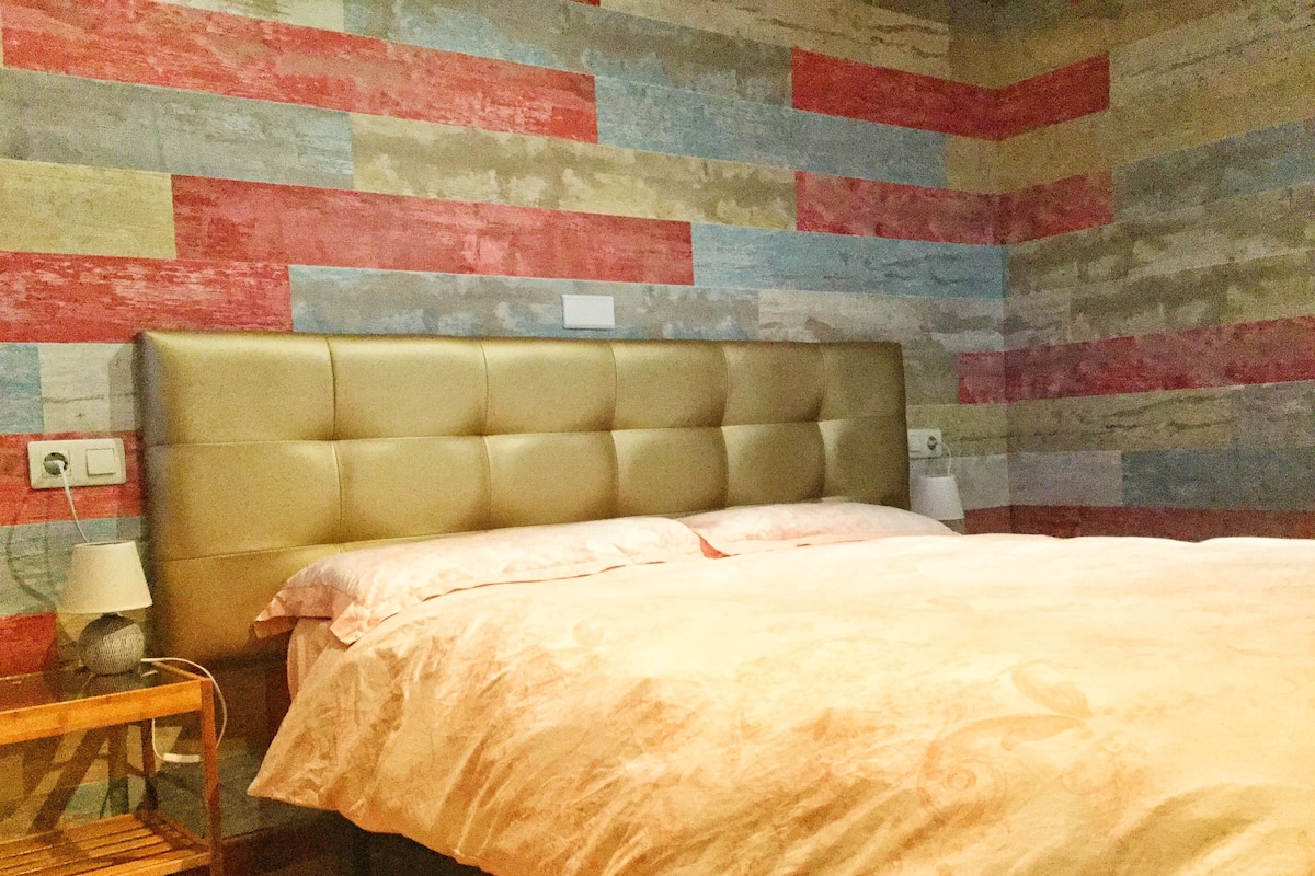 A bedroom with a tufted headboard, a bed with a light-colored duvet, a bedside table with a lamp, and a wall with a multicolored striped pattern.