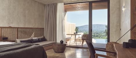 A modern bedroom with a large bed, a wooden desk, a chair, and a view of a mountainous landscape through a sliding glass door.