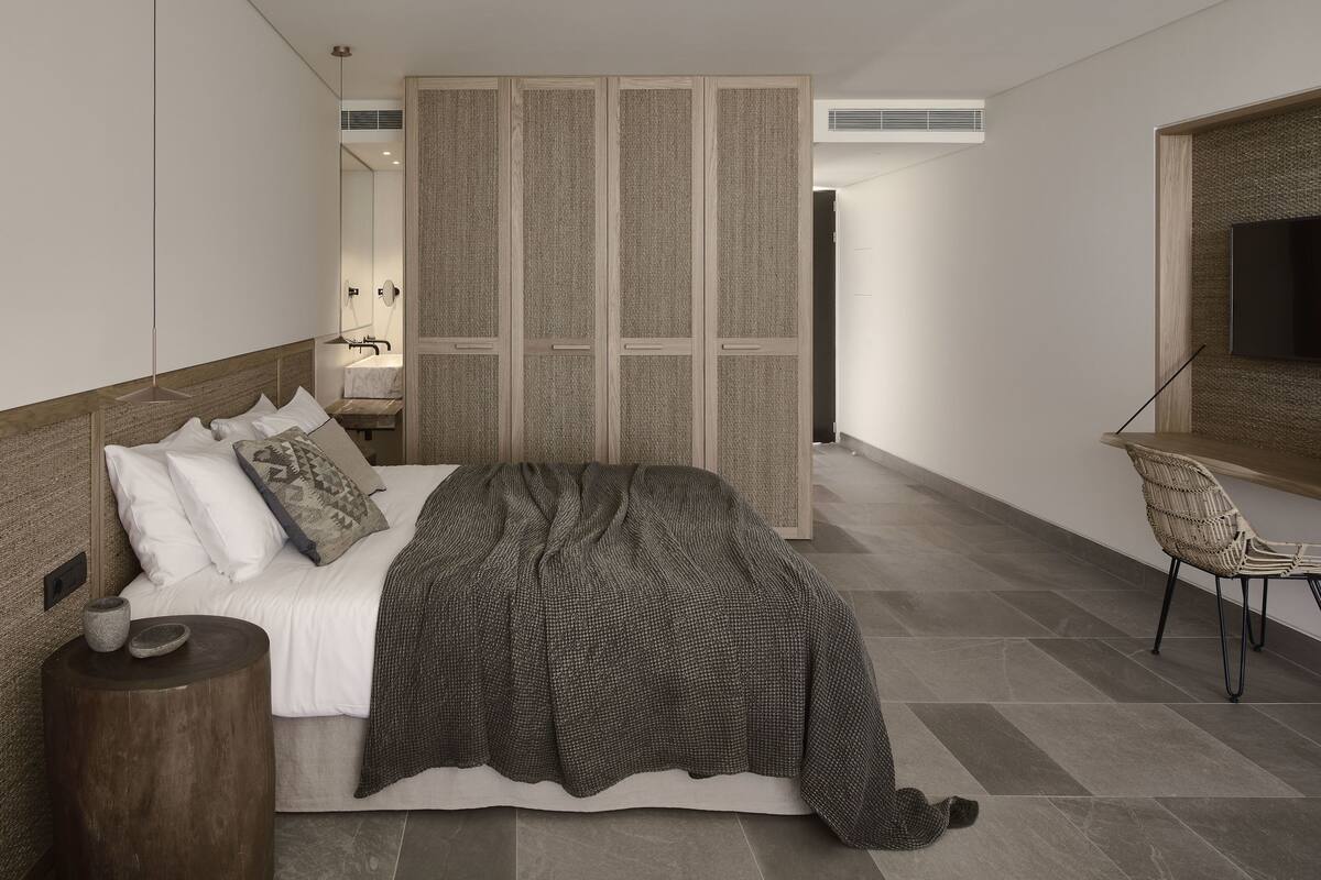 A modern bedroom with a large bed, a wooden headboard, a bedside table with a lamp, a woven chair, and a large window with a view of the city.