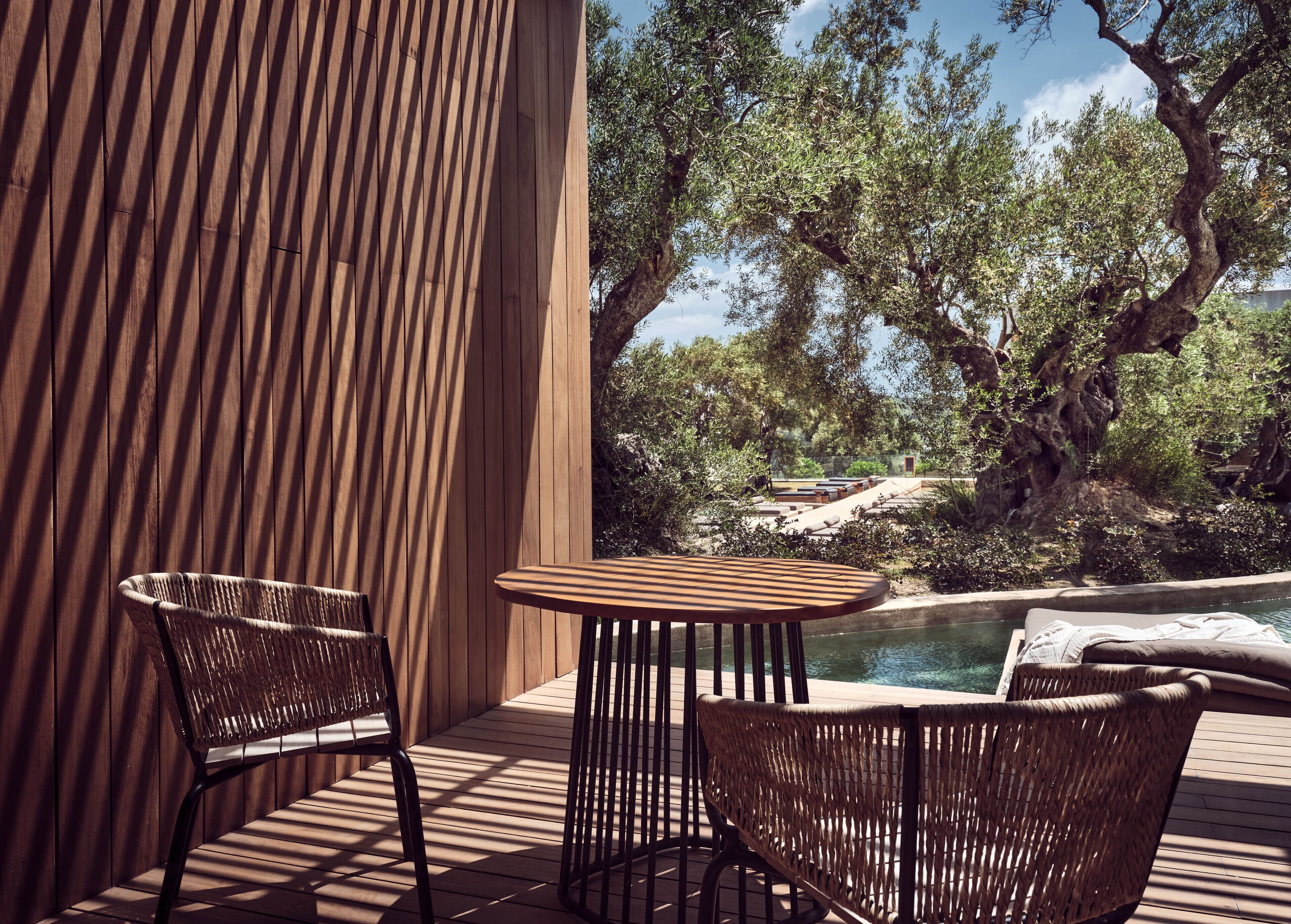 A wooden deck with two wicker chairs and a small table, overlooking a pool and natural landscape.