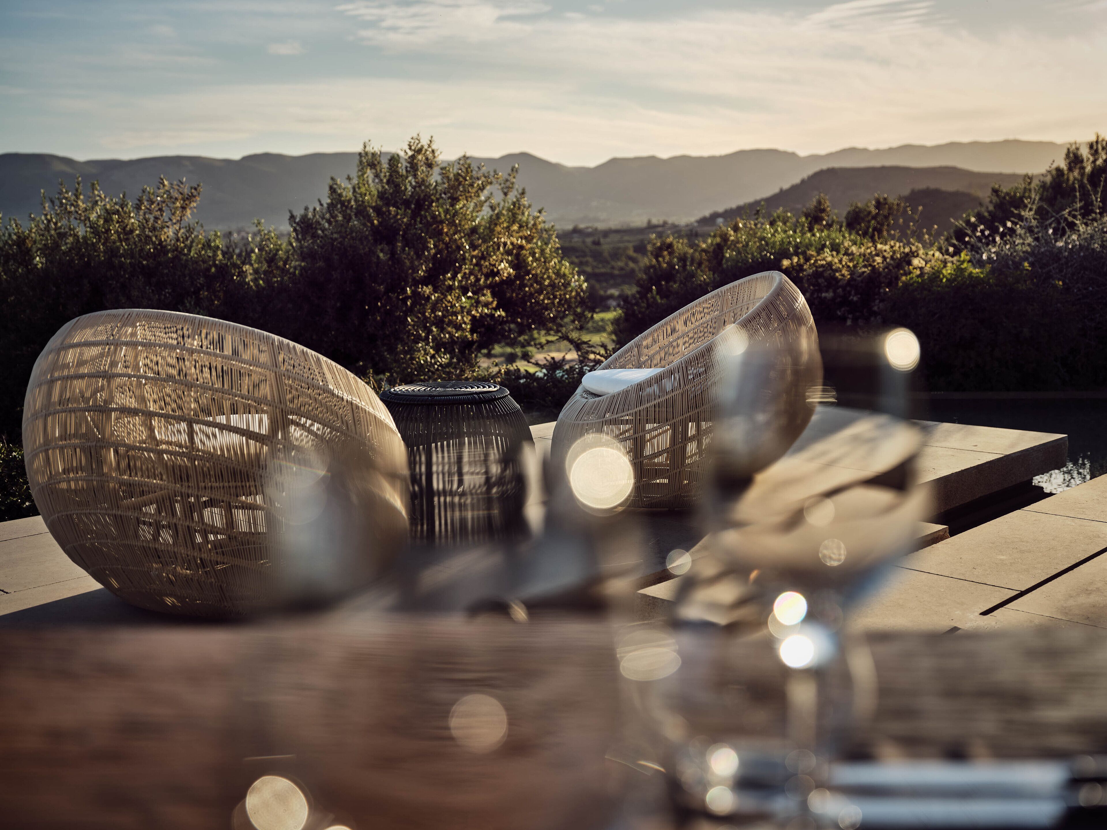 A dining area with a view of mountains and a body of water. There are two large woven chairs and a table with glasses.