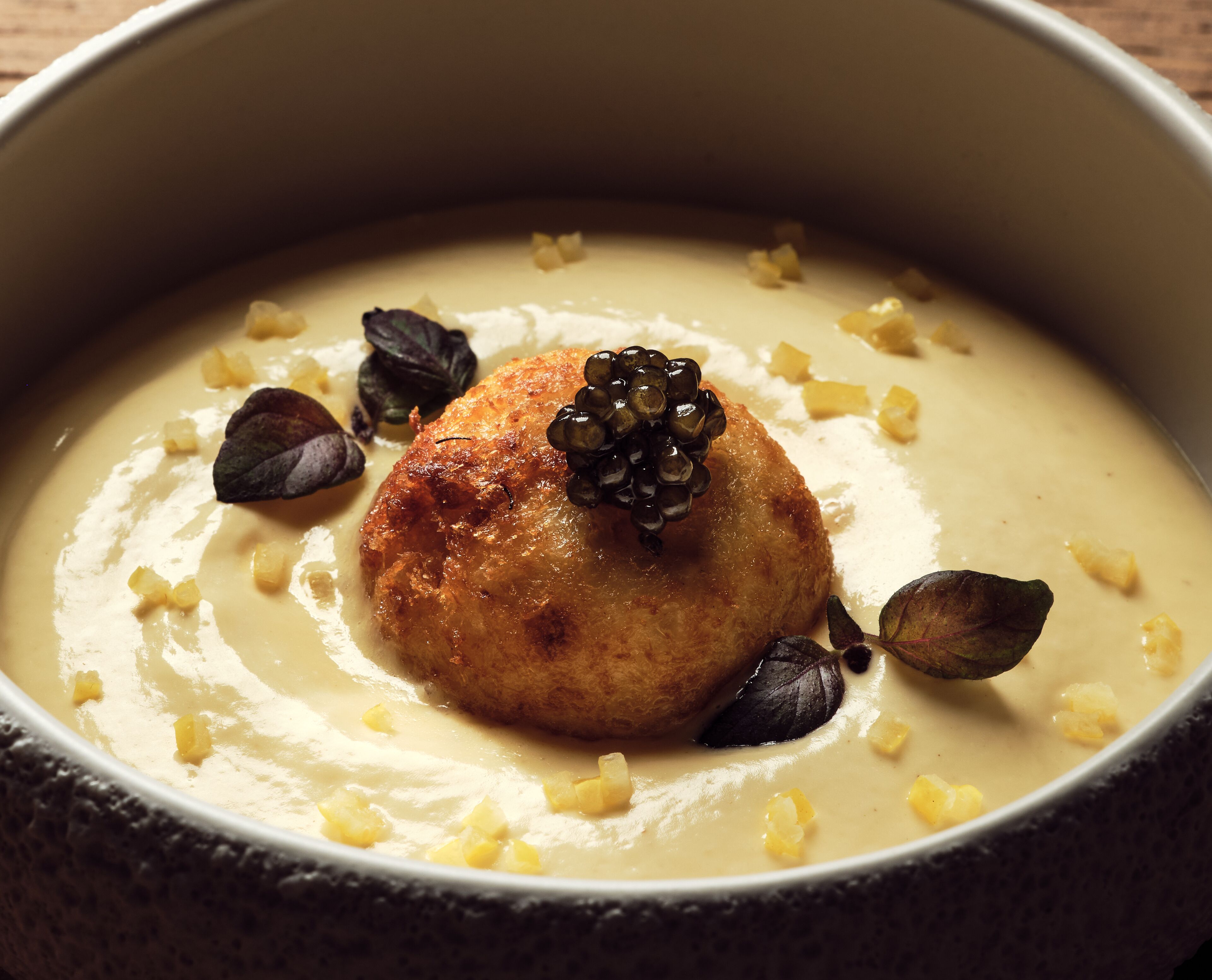 A bowl of creamy soup with a round, golden-brown item on top, garnished with small cubes and dark leaves.