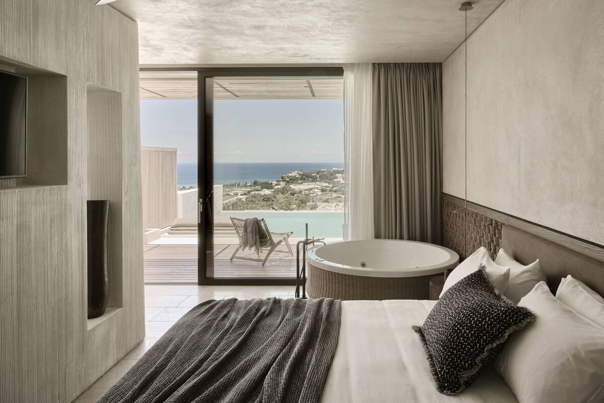 A modern hotel room with a large bed, a round bathtub, a TV, and a balcony with a view of the sea.