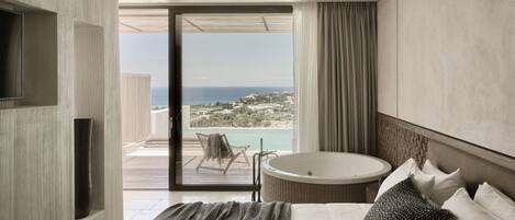 A modern hotel room with a large bed, a round bathtub, and a balcony with a view of the sea.