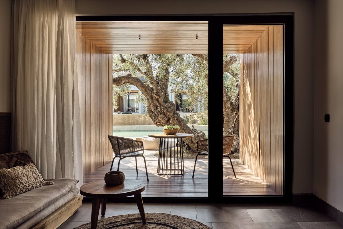 A modern living room with a sofa, a round table, and two chairs. A large tree is visible through the sliding glass door, and a pool is seen in the background.
