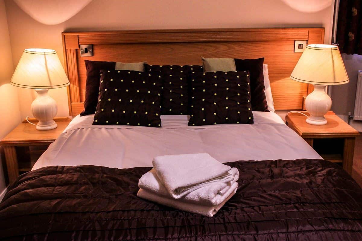 A neatly made bed with a dark brown comforter, two white lamps on either side, and a wooden headboard.