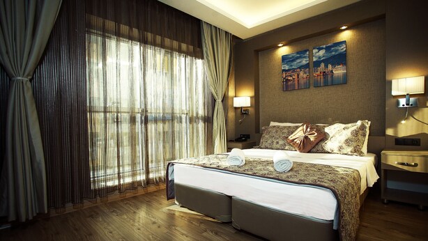 A hotel room with a large bed, a nightstand, a lamp, and a cityscape painting on the wall. - Hotel Iz (Izmir)