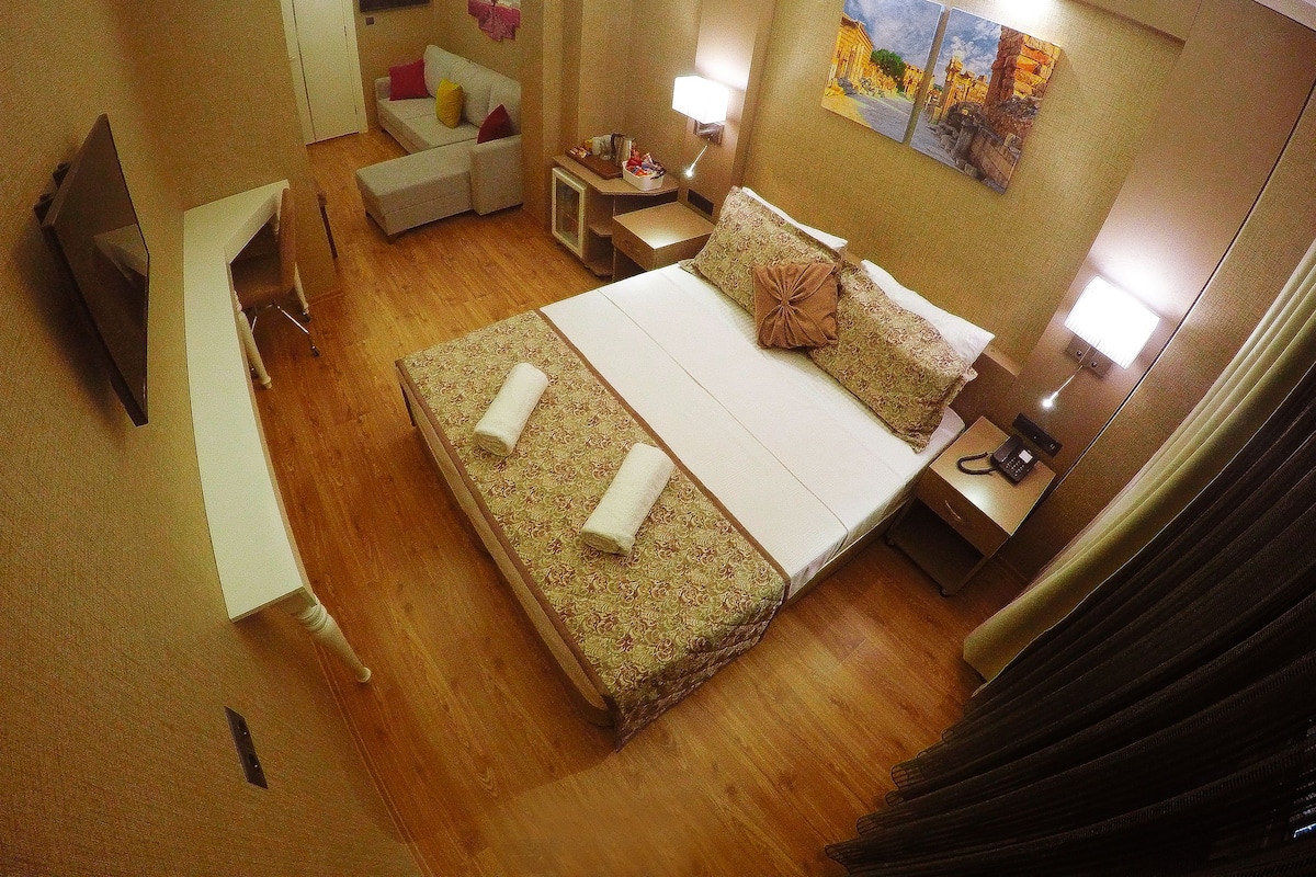 A hotel room with a large bed, a desk, a chair, a TV, and a small table with a lamp.
