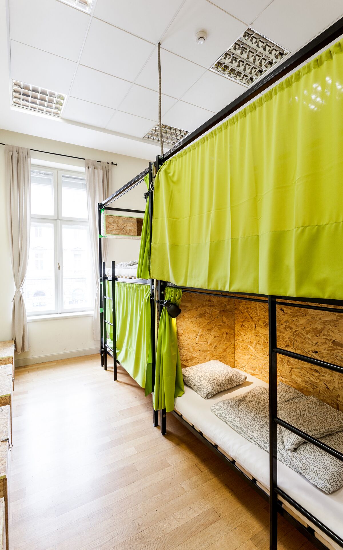 A dormitory room with bunk beds, wooden flooring, and a window with curtains.