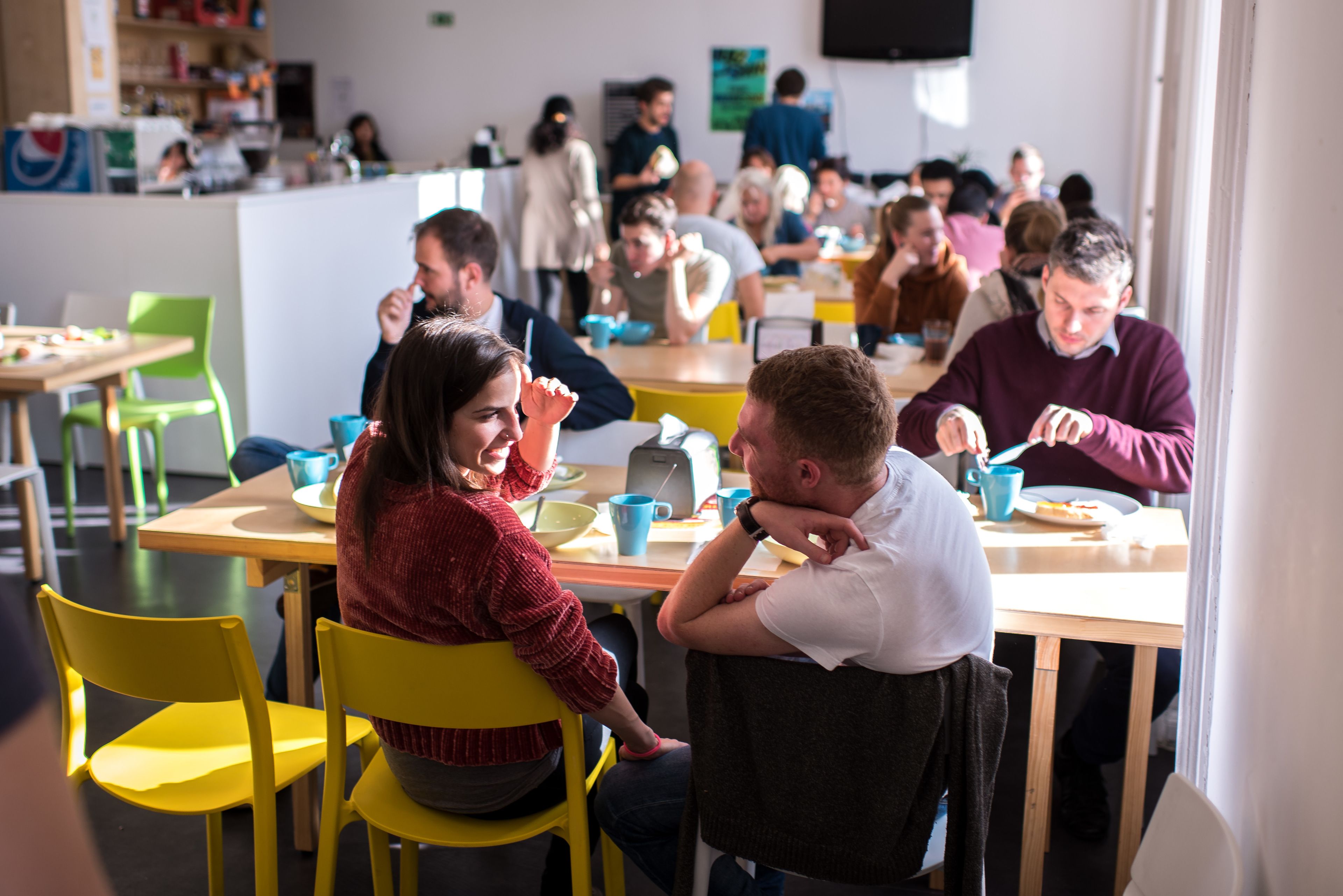 Un comedor compartido con sillas y mesas amarillas, una barra con alimentos, y un grupo de personas conversando.