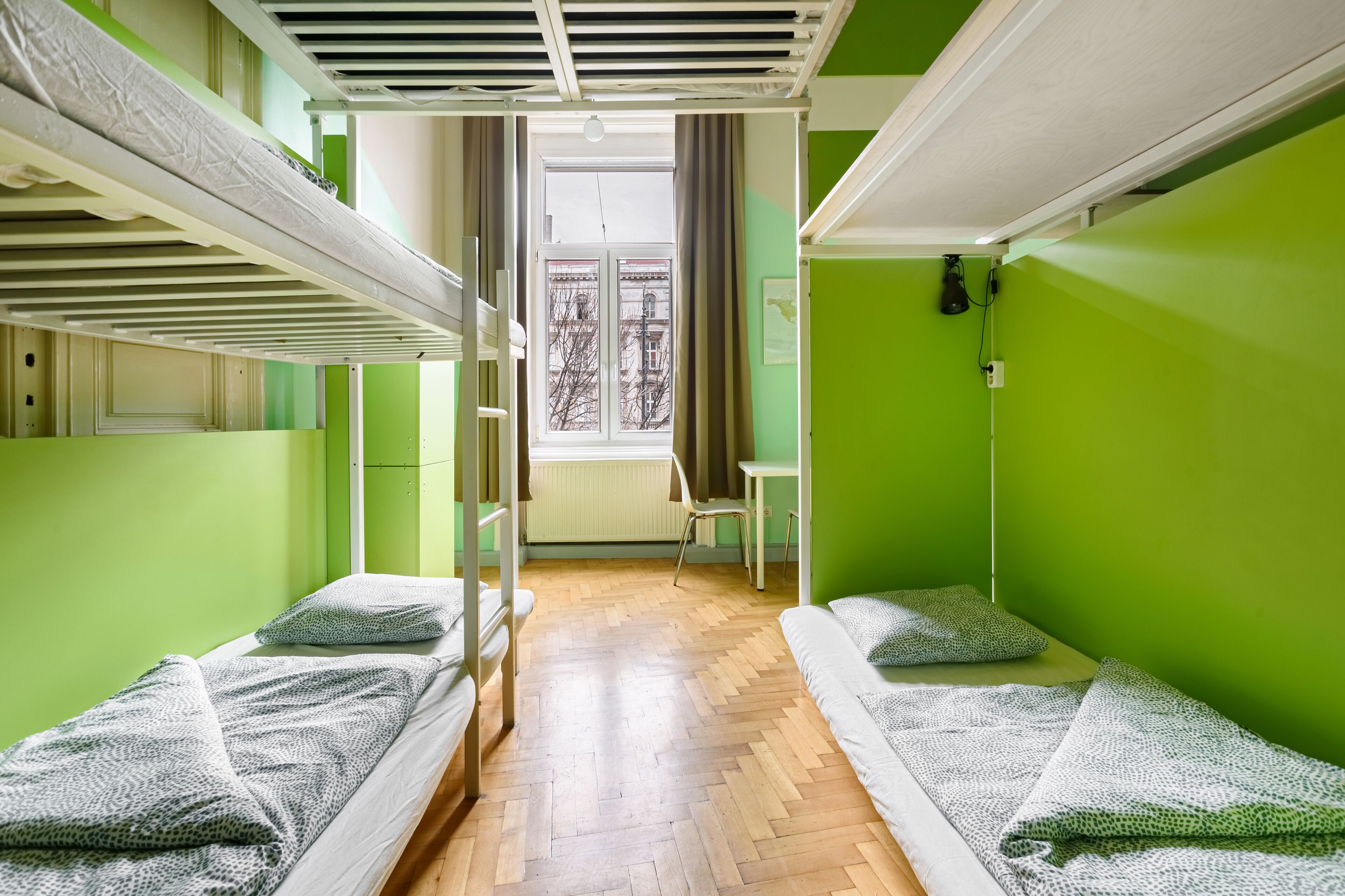 A dormitory room with bunk beds, a wooden floor, a window with a view of buildings, and a radiator.