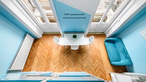 A room with a herringbone wooden floor, a blue sofa, a white table with a potted plant, and a ceiling sign displaying a WiFi password. - Avenue Hostel (Budapest)