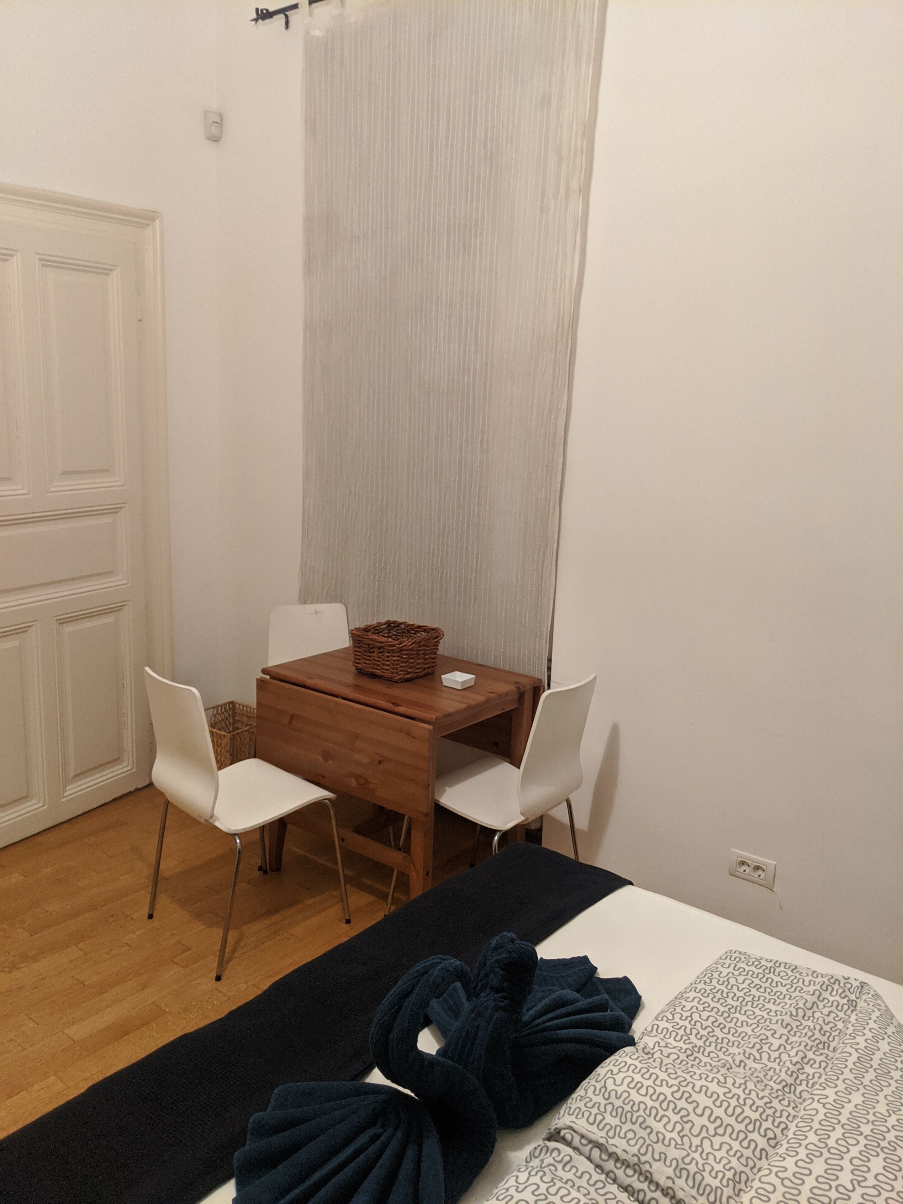 A small, tidy room with a wooden table and two white chairs, a bed with a dark blanket, and a white door.