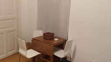 A small, tidy room with a wooden table and two white chairs, a bed with a dark blanket, and a white door.
