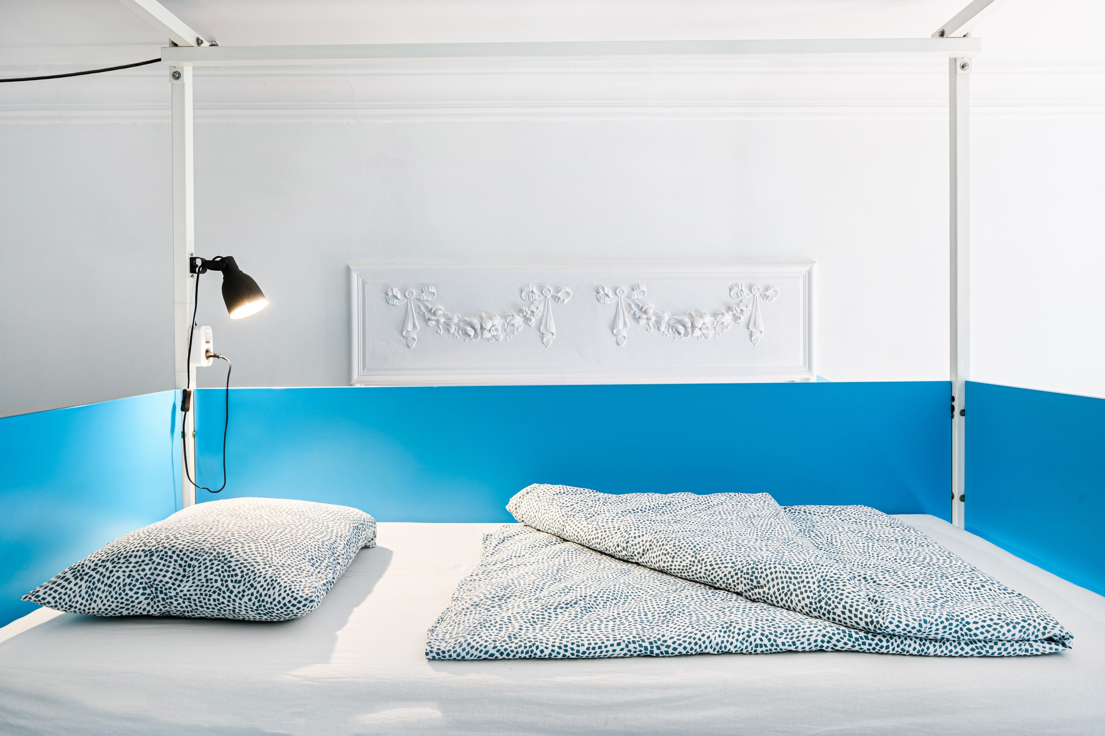 A bed with a white and blue color scheme, a wall-mounted lamp, and a decorative piece above the bed.