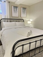Egy gondosan megterített ágy fehér ágyneművel és párnákkal, fekete fémvázas ágykerettel, és egy feltekert törölközővel az ágyon. A falon, az ágy felett két kép van keretben, az éjjeliszekrényen pedig egy asztali lámpa.