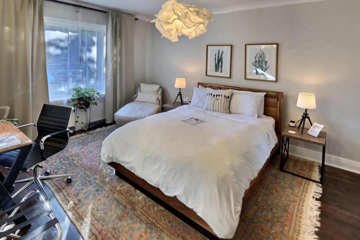 Habitación de hotel con una cama grande, dos mesitas de noche con lámparas, un escritorio con una silla, un ventanal con cortinas y dos obras de arte en la pared.