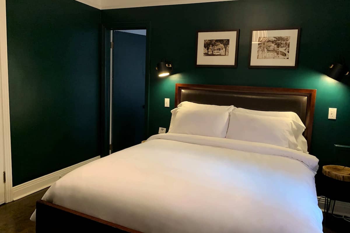 Una cama bien hecha en una habitación con paredes verde oscuro, dos cuadros en la pared y una mesita redonda con una lámpara junto a ella.
