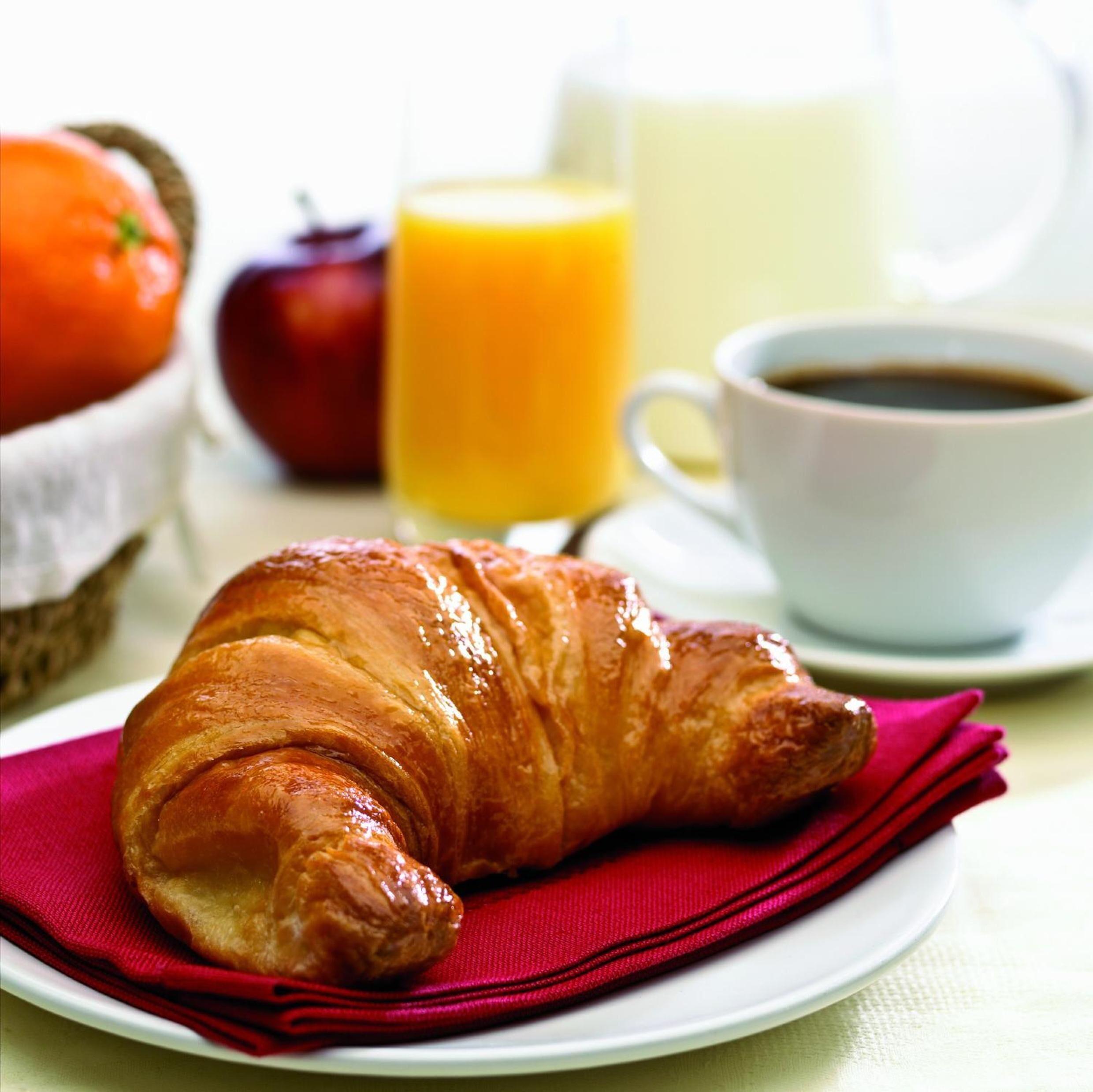 A croissant on a plate with a red napkin, a glass of orange juice, a cup of coffee, and a basket with an apple and a tomato.