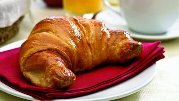 A croissant on a plate with a red napkin, a glass of orange juice, a cup of coffee, and a basket with an apple and a tomato.