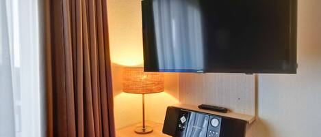 Habitación de hotel con televisor de pantalla plana montado en la pared, mesita de noche con lámpara y control remoto.