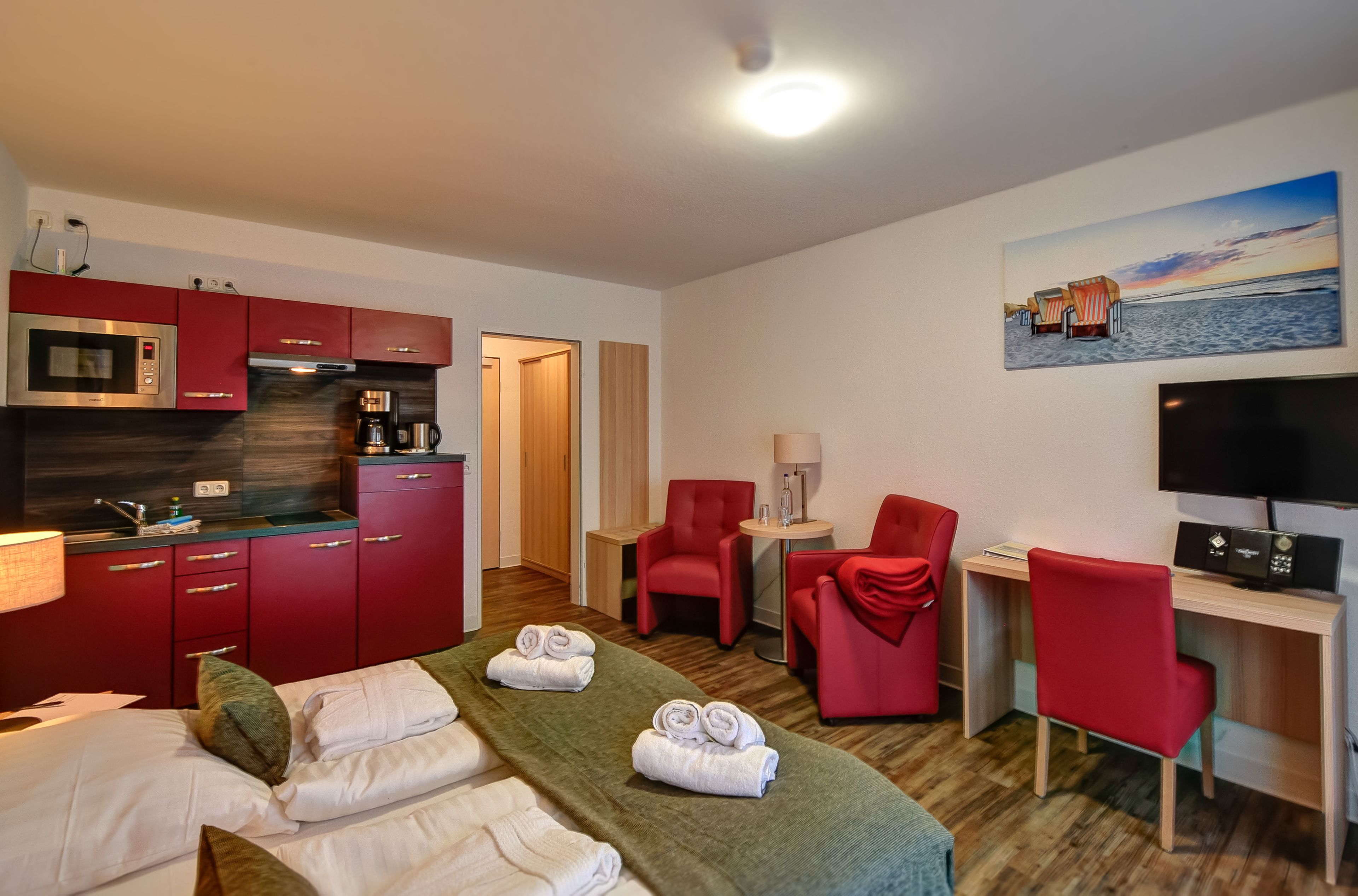 Une chambre d’hôtel avec un lit, un bureau, une télévision, une chaise rouge, un fauteuil rouge, une machine à café et un tableau représentant une scène de plage.