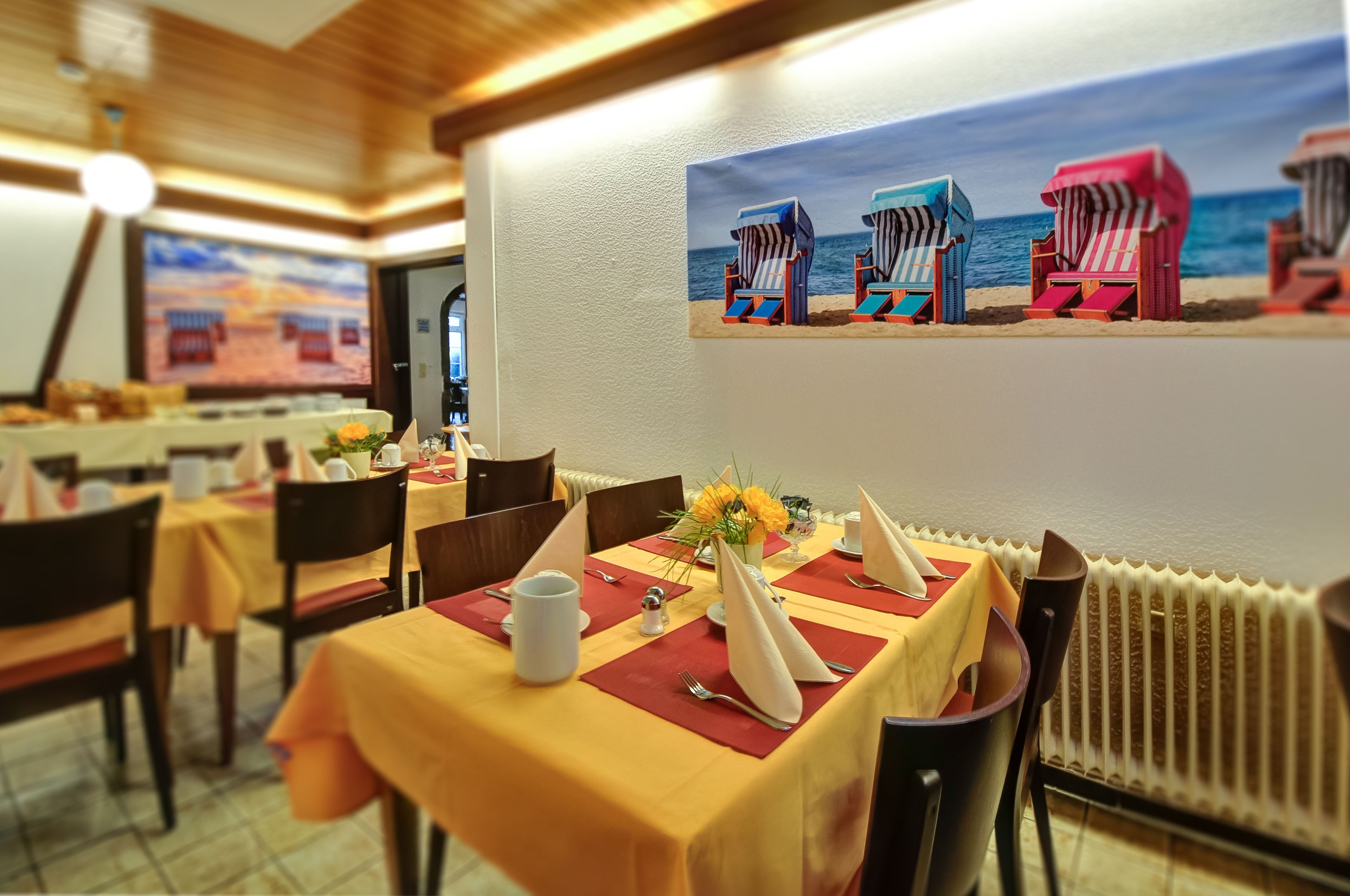 A dining area with tables set for two, featuring yellow tablecloths and red placemats. A painting of beach chairs is on the wall.